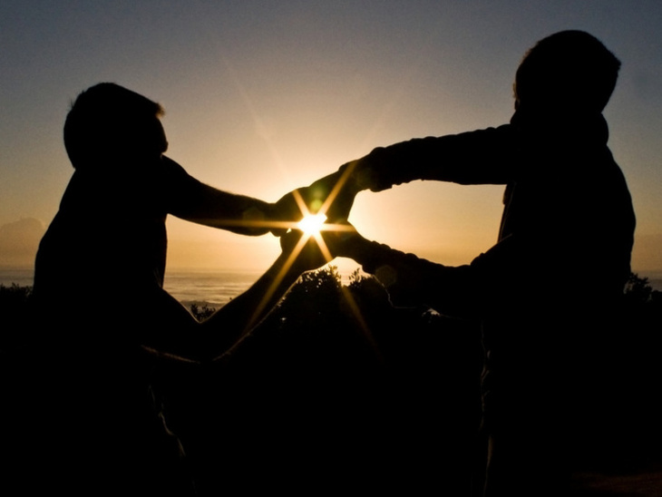 Zwei Personen als Silhouetten vor einem Sonnenuntergang, die sich an beiden Händen fassen – die untergehende Sonne scheint genau zwischen ihren berührenden Händen hindurch und erzeugt einen Lichtstrahl.