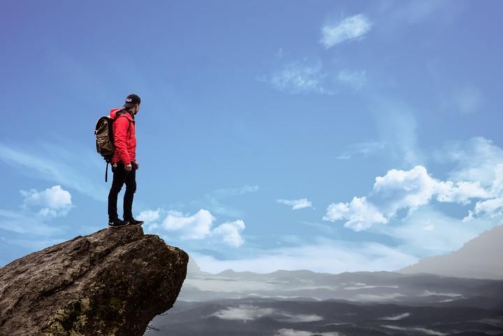Person mit roter Jacke und Rucksack steht auf einem Felsen und blickt über eine weite Berglandschaft unter blauem Himmel.